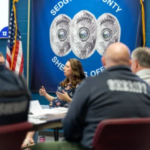 A bald Sheriff-jacketed deputy listens from behind as trainer Sarah Thome presents before the Sedgwick County banner at the Wichita workshop.