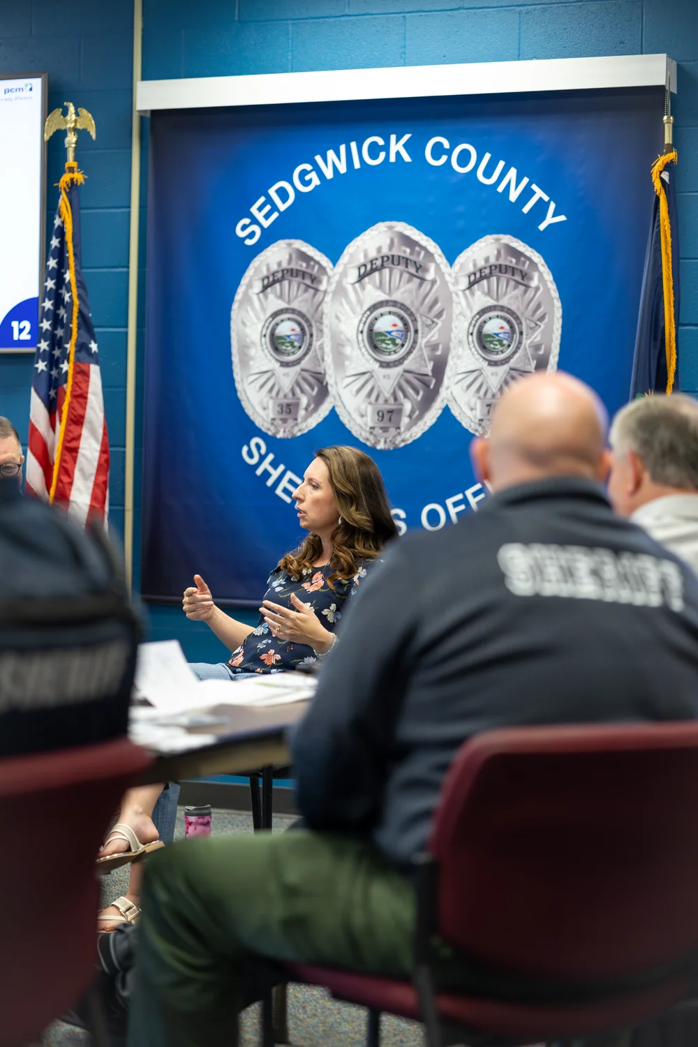 A bald Sheriff-jacketed deputy listens from behind as trainer Sarah Thome presents before the Sedgwick County banner at the Wichita workshop.