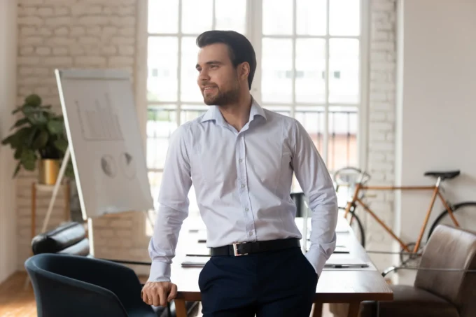 A bearded man in a light dress shirt stands smiling by a desk in a loft office with a flip chart.