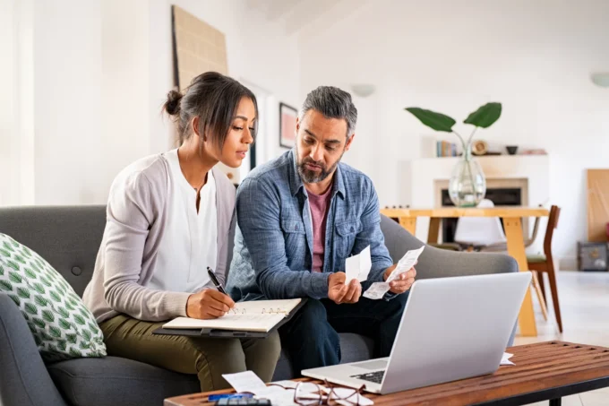 Man and woman sit on a sofa reviewing receipts and taking notes with a laptop while managing home finances