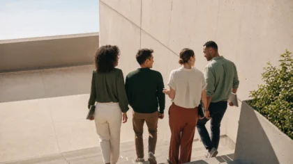 From behind, a group of multiracial colleagues walking side by side down an exterior concrete stairwell carrying laptops and folders