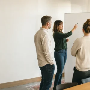 A group of white / Caucasian colleagues gathered standing around a whiteboard mid-discussion, one gesturing at the board while others lean