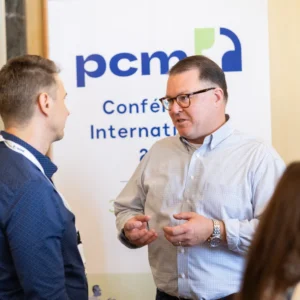 Sarah Thome gestures with both hands while conversing with a woman beside a wooden table at the Bordeaux PCM® conference.