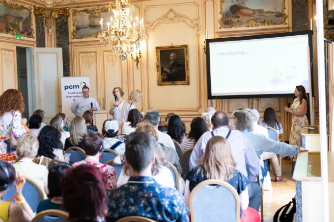 Three speakers present the PCM® Relationship Profile to a large seated audience under a chandelier at the PCM® International Conference in Bordeaux.