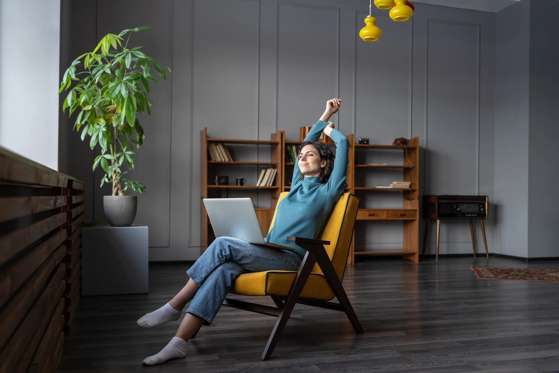 Young woman stretching contentedly with a laptop on her lap while relaxing in a yellow armchair at home.