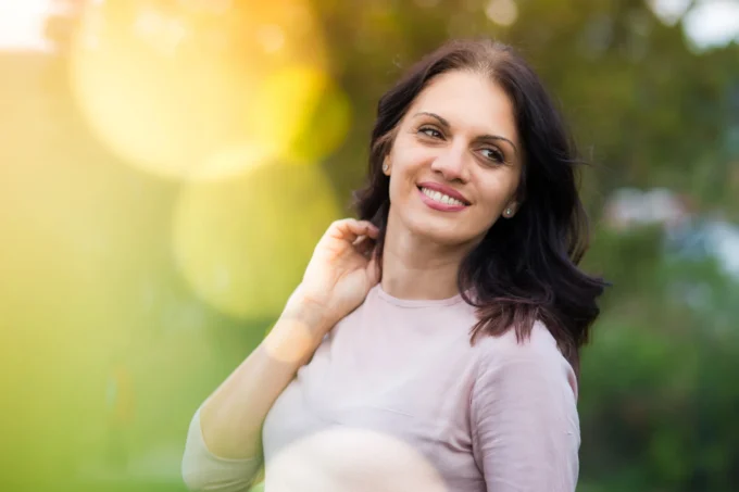 A dark-haired middle-aged woman smiles and gazes away thoughtfully in a sunlit outdoor portrait with green background
