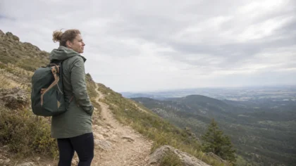Wide environmental shot of a white / Caucasian person paused partway up a switchback hiking trail, looking out over the ridge; overcast midday