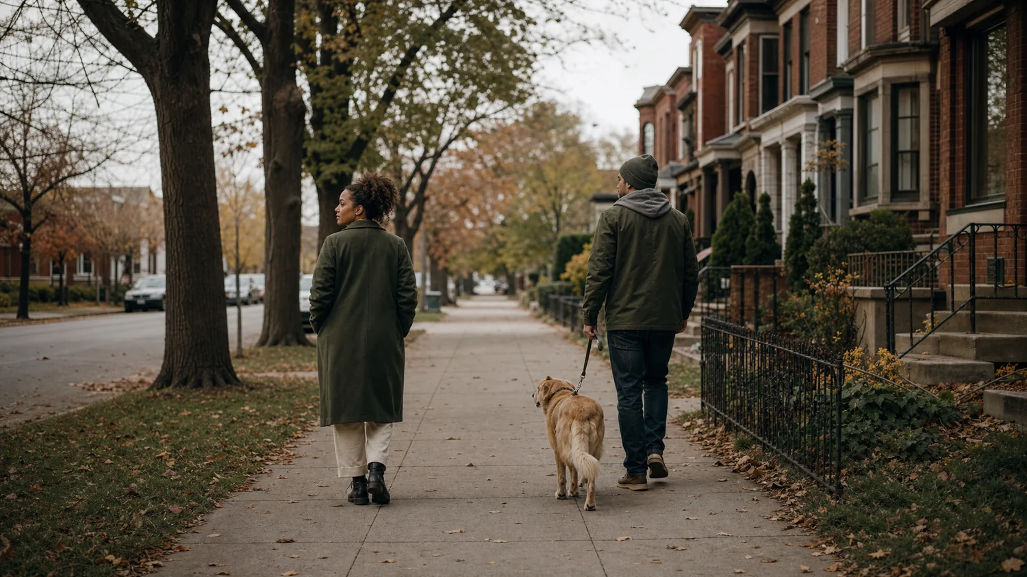 A multiracial couple (one woman, one man) walking a dog along a tree-lined sidewalk with a small gap between them, one gazing off to the side