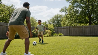 A Black / African American parent and their young child kicking a soccer ball back and forth in a grassy backyard