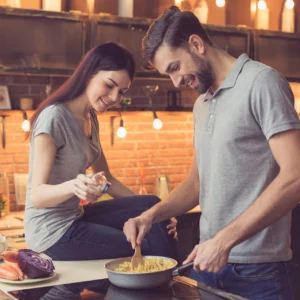 A smiling young couple prepares food together at a stovetop in a warmly lit brick kitchen.