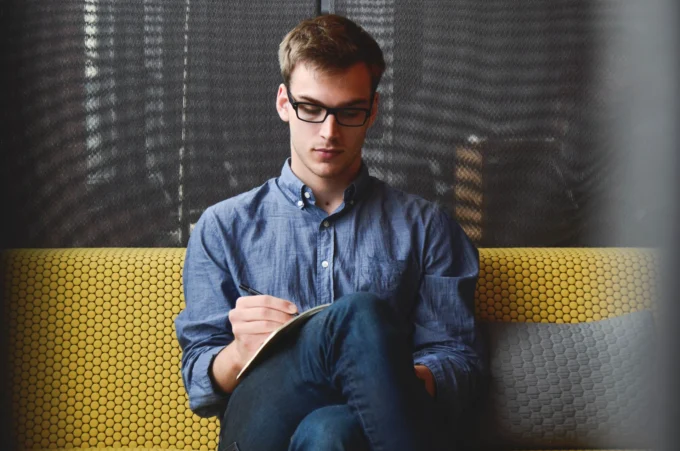 Focused young man in glasses and a blue shirt writes in a notebook while sitting on a yellow bench.
