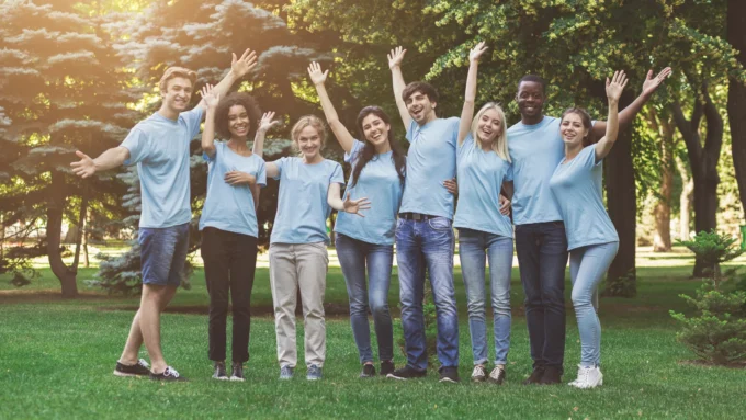 Diverse group of eight young people in matching blue t-shirts raise their arms and smile together in a park